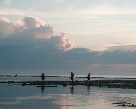 An image of a tranquil beach during sunset. The sky displays hues of pink and purple, with large fluffy clouds in the distance. The calm water reflects the sky, creating a mirrored effect. Silhouettes of three individuals can be seen walking and standing on the wet shoreline, contributing to the calm and reflective ambiance of the scene