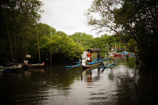 A traditional Balinese jukung boat with a thatched roof, floating gently on the calm waters among the mangroves, with passengers enjoying the natural shade and scenery.