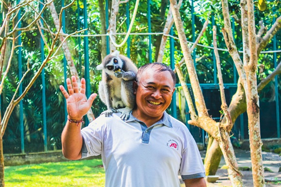 A smiling man with a friendly lemur perched on his shoulder, set against the lush greenery of a wildlife sanctuary