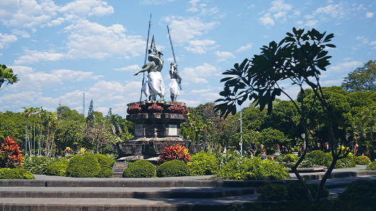 The image shows a monument featuring three statues holding spears, wrapped in traditional white cloths, standing on a raised stone pedestal. The monument is set within a beautifully landscaped park filled with tropical plants, neatly trimmed bushes, and vibrant flowers. A clear blue sky with light clouds provides a bright backdrop to the scene. The statues appear to commemorate a significant historical or cultural event, while the surrounding greenery creates a peaceful and inviting environment for visitors. The overall setting conveys a sense of respect, tradition, and tranquility.