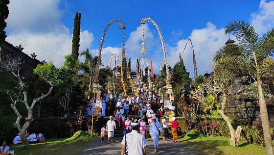 This image shows the entrance to Besakih Temple, often referred to as the "Mother Temple" of Bali. Worshippers dressed in white are seen ascending the temple steps, while the entrance is decorated with tall penjors, bamboo poles traditionally used in ceremonies, adding to the sacred and festive atmosphere of this major spiritual site.