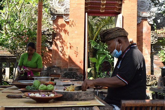 Balinese cooking instructor preparing fresh ingredients in an outdoor kitchen, surrounded by traditional architecture and greenery, showcasing the authentic culinary experience of learning to cook local dishes in Bali