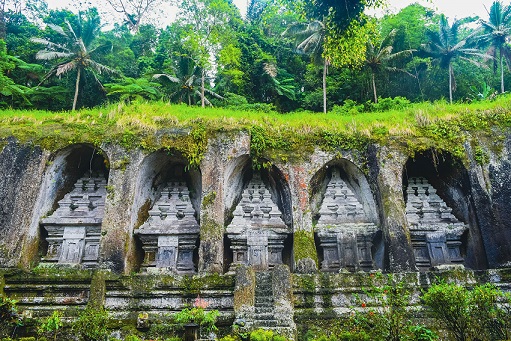 The image shows a row of ancient stone shrines, or candi, carved into the cliffside at Gunung Kawi Temple. The shrines are nestled under a lush green hill, surrounded by tropical palm trees and dense vegetation, giving the temple a peaceful and natural atmosphere. The intricate carvings in the rock contrast beautifully with the vibrant greenery above, creating a striking visual of Bali’s ancient culture blended with nature. This sacred site reflects Bali’s historical and spiritual significance, making it a remarkable destination for visitors.