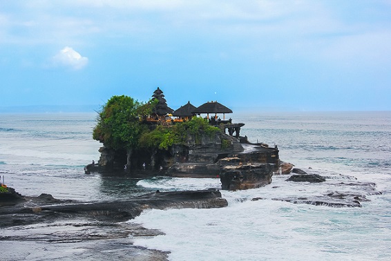 The image shows Tanah Lot Temple sitting majestically on a rocky island, surrounded by the ocean. The temple, with its traditional Balinese architecture, stands out against the backdrop of the vast, calm sea. Waves gently lap against the base of the rock, while lush greenery adorns the temple's surroundings. The sky is clear with a soft blue hue, complementing the serene atmosphere of this iconic coastal temple.