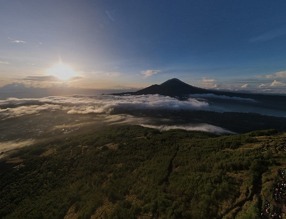 A breathtaking view of Mount Batur at sunrise, surrounded by layers of clouds and green hills. The sun rises over the horizon, casting a warm glow across the landscape and illuminating the valleys and forested areas below. The scene captures the natural beauty and tranquility of Kintamani from above, showcasing the majestic appeal of Bali's volcanic highlands.