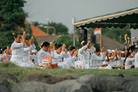 A group of Balinese women dressed in white participating in a prayer ceremony, sitting with their hands together in a gesture of worship, symbolizing devotion and respect.