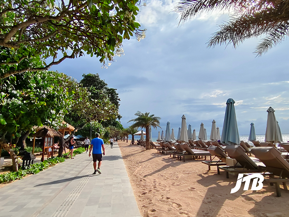 A scenic view of Sanur Beach boardwalk in Bali, featuring a paved pathway lined with lush greenery, sun loungers, and beach umbrellas