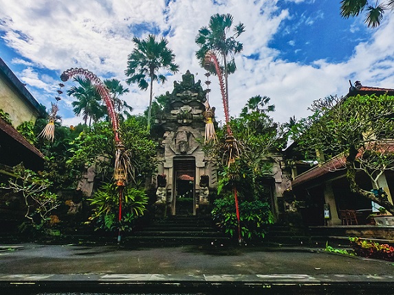 Ornate entrance to a traditional Balinese building surrounded by lush greenery and tall palm trees under a partly cloudy sky, highlighting Bali's cultural architecture