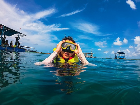 Person snorkeling in clear blue waters with a mask and snorkel, surrounded by a bright sky and boats in the background