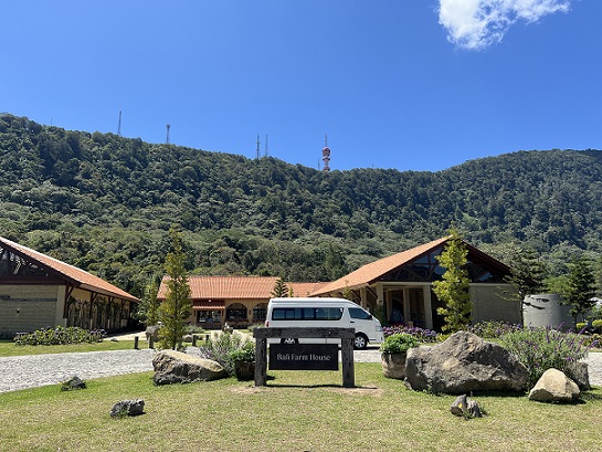 Entrance to Bali Farm House with traditional countryside-style buildings set against a backdrop of lush green hills under a bright blue sky