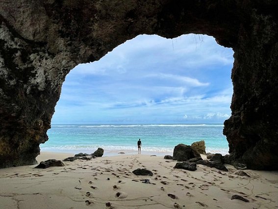 A person stands on a sandy beach framed by a large rocky cave opening, with clear turquoise waters and a blue sky in the background