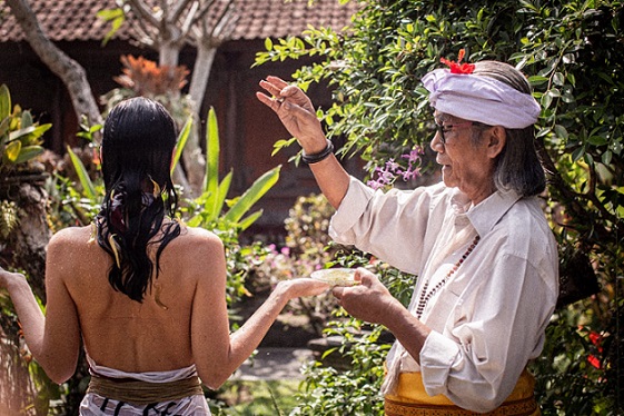 A Balinese healer performs a traditional blessing ritual on a visitor, sprinkling holy water in a serene garden setting