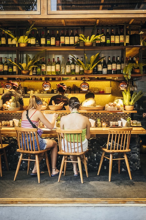 Two women dining at a cozy, rustic eatery with a bar-style counter. The setting features warm lighting, wooden chairs, and a backdrop filled with wine bottles and greenery, creating a relaxed, intimate atmosphere