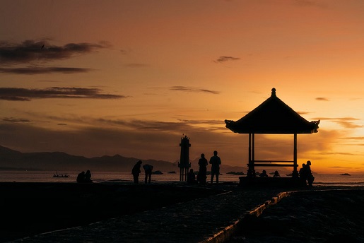 Silhouettes of visitors enjoying a serene sunrise at Sanur Beach, Bali, with a traditional gazebo in the foreground