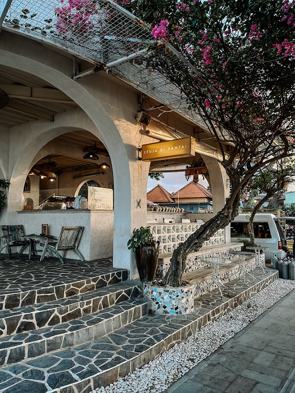 Exterior view of Stuja di Pantai café in Sanur, showcasing its minimalist, open-air design with stone steps, arched entryways, and vibrant bougainvillea flowers enhancing the aesthetic. The café exudes a cozy, beachfront vibe, perfect for relaxing by the ocean