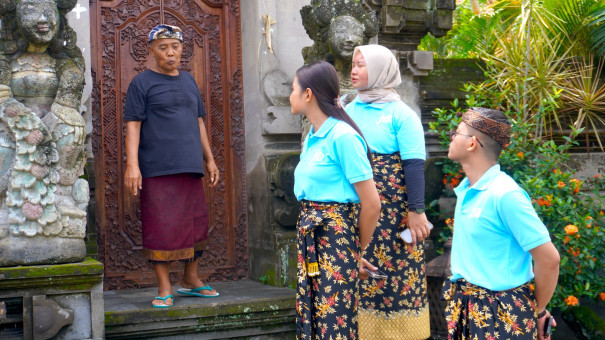 Visitors in traditional Balinese attire engaging in a cultural exchange with a local elder at the entrance of a traditional Balinese home, surrounded by intricate stone carvings and lush greenery.