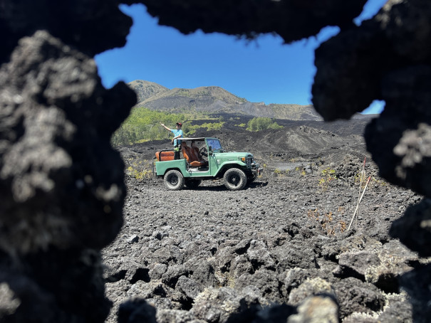 A vintage off-road Jeep navigating the rugged black lava fields of Mount Batur in Bali, framed by volcanic rock formations. The adventure tour offers a unique experience exploring the dramatic volcanic landscape.