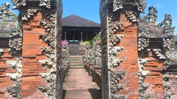 Traditional Balinese red-brick temple gate with intricate stone carvings, leading to a pavilion flanked by guardian statues and steps—part of a heritage site under clear blue sky.