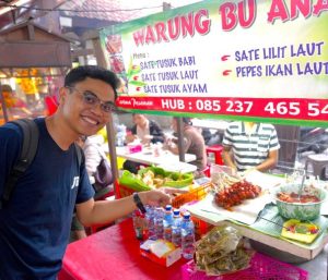 Meanwhile, an evening Bali street food scene in Denpasar shows a local satay stall with grilled skewers, banana leaf–wrapped dishes, and a traveler pointing at the food counter.
