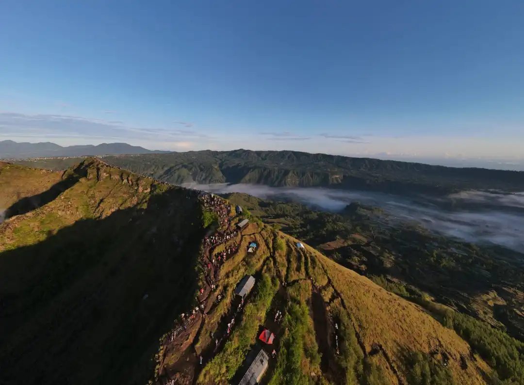 Aerial view of a hiking trail on Mount Batur in Bali, Indonesia, with a line of trekkers visible against the mountain's ridge. The landscape is bathed in the warm glow of morning light, with mist-filled valleys stretching into the distance and the caldera's edge defining the horizon