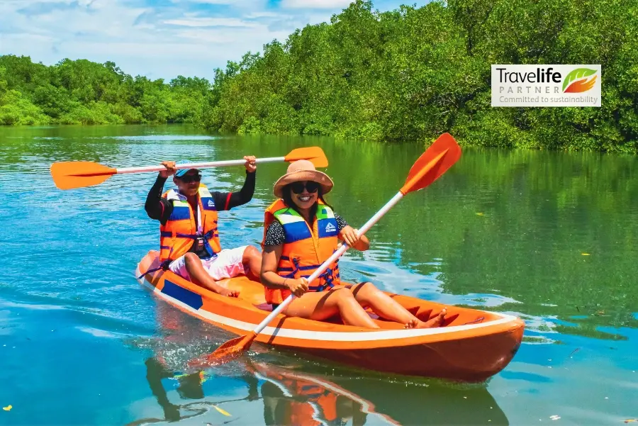 Two individuals in orange kayaks on a calm river surrounded by lush greenery. They wear bright orange life vests and sun hats, paddling with orange oars. In the top right corner, there's a logo for 