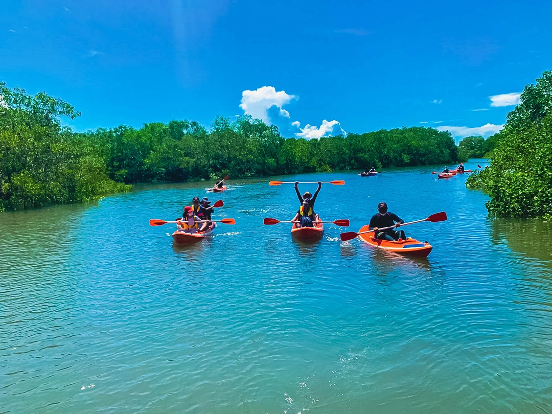 Several individuals paddle in bright orange kayaks on a serene blue waterway, surrounded by lush green mangrove trees under a clear blue sky. The forefront features a pair in a tandem kayak, while others are spread out, enjoying the tranquil environment. The reflections in the water add to the picturesque nature of the scene