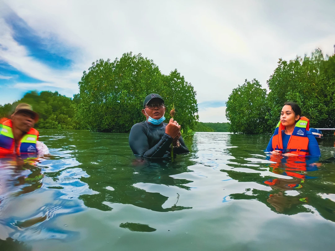 Three individuals wearing life vests are waist-deep in clear water, surrounded by lush mangroves. The central person, wearing glasses and a cap, appears to be talking or giving instructions, while a woman to the right listens attentively. The leftmost individual is slightly blurred in motion. Their reflections ripple on the water's surface, and the sky above is overcast