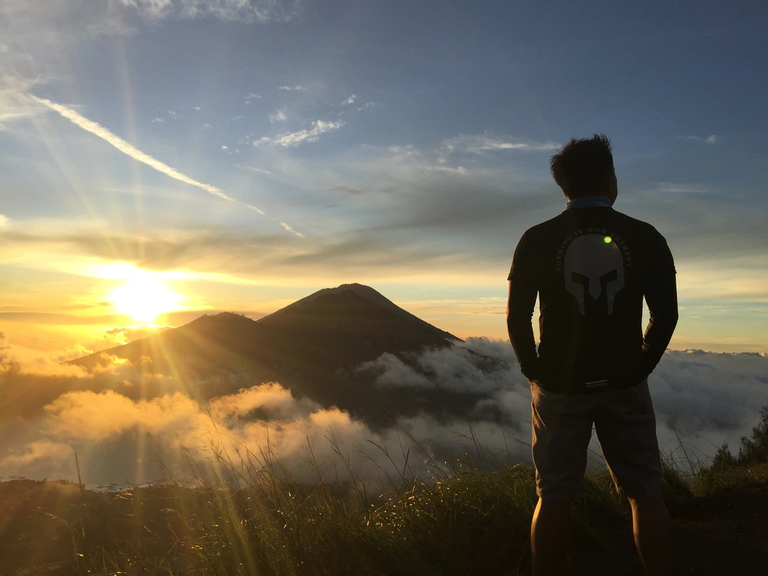 A silhouette of a person standing at the summit, gazing at the sunrise over Mount Batur in Bali, Indonesia. The early morning sky is painted with hues of orange and blue, while clouds envelop the lower landscape, creating a breathtaking view from the mountaintop