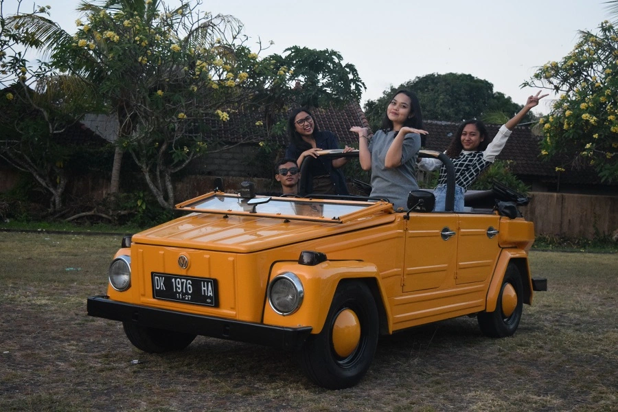 A group of four people, three women and one man, joyfully posing in an orange vintage Volkswagen Thing car, parked on grass with frangipani trees around.