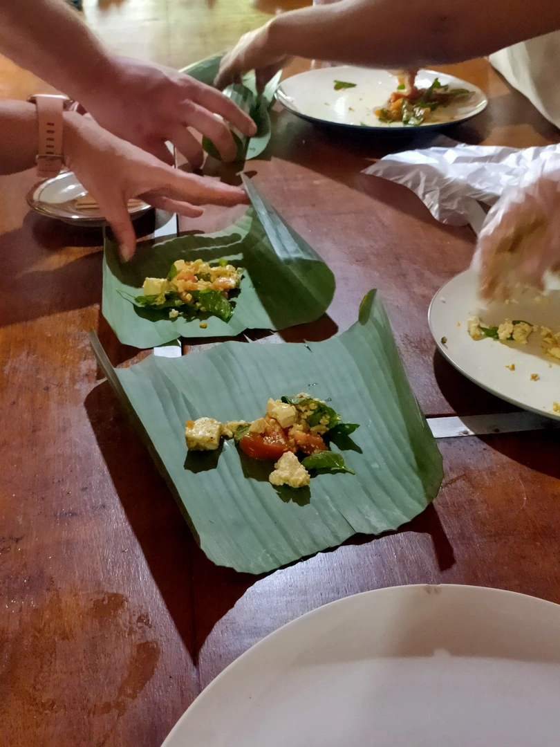Participants in a cooking class prepare dishes on a wooden table, using banana leaves as natural plates. They are assembling ingredients for a traditional Balinese meal, with hands visible working on the dishes, and wearing gloves for hygiene