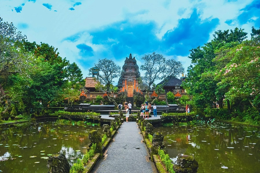 Photograph of the Puri Saraswati, also known as Saraswati Temple, in Bali, Indonesia, viewed from across a pond filled with lotuses. A path leads to the temple, flanked by lush greenery, with the temple's ornate, multi-tiered meru towers rising against a blue sky.