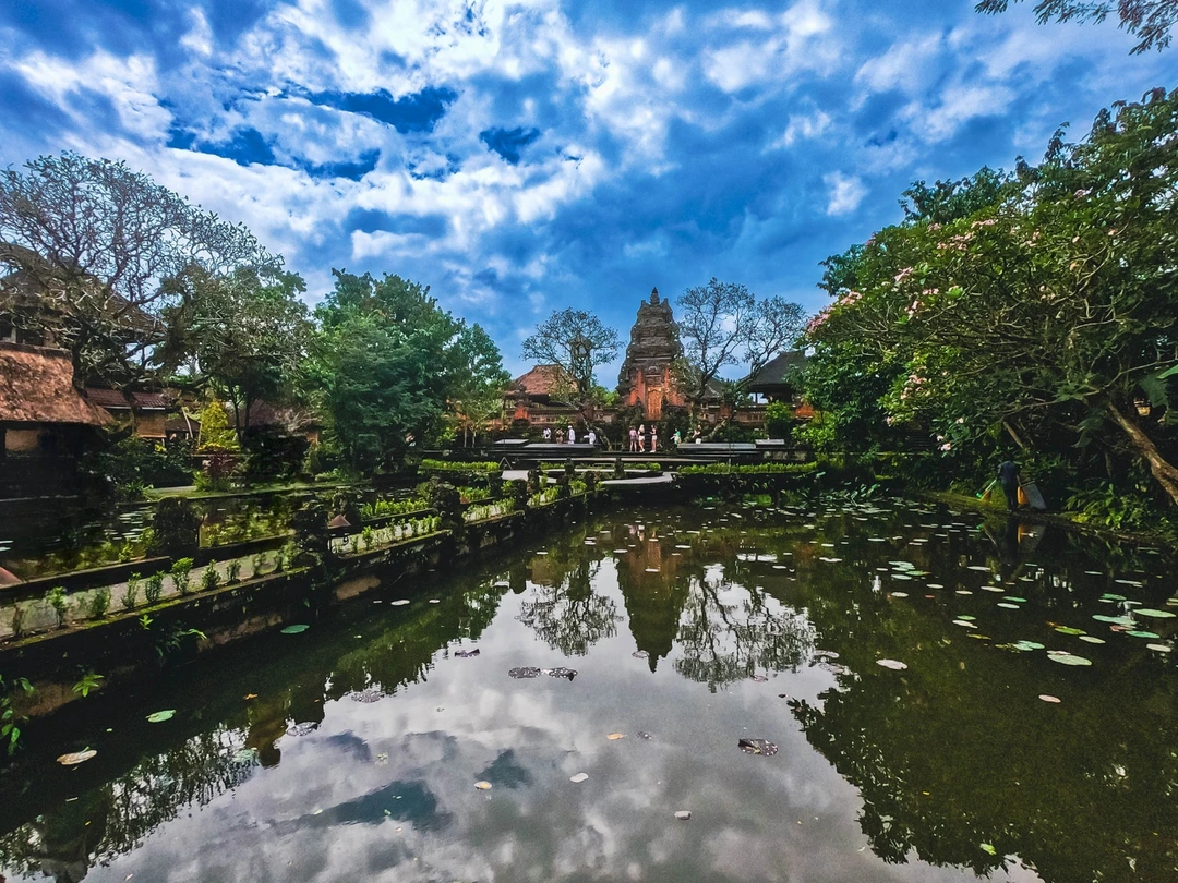 Expansive view of the Puri Saraswati Temple in Bali, Indonesia, with its reflection mirrored in a tranquil foreground pond dotted with water lilies. Dramatic clouds scatter across the sky, enhancing the temple's serene and majestic presence amidst lush tropical foliage