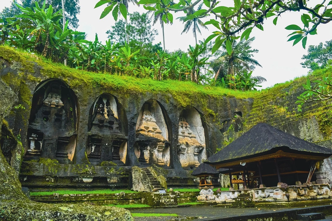 Image of the ancient Gunung Kawi Temple in Bali, Indonesia, showing a series of large, carved stone shrines set into a moss-covered rock face. A traditional thatched-roof pavilion stands in the foreground, with lush tropical vegetation and tall palm trees in the background, creating a serene and historic atmosphere
