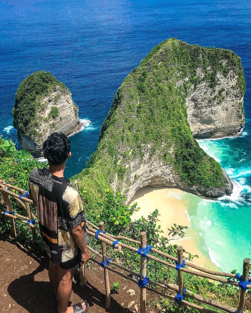 A traveler stands at a viewpoint overlooking the iconic Kelingking Beach on Nusa Penida Island, Bali. The viewpoint reveals a dramatic, T-rex-shaped cliffside plunging into turquoise waters, with a secluded white sand beach nestled at its base