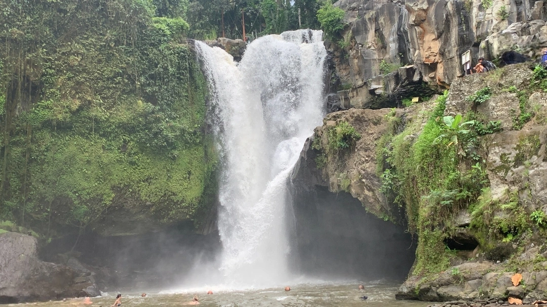 The powerful Tegenungan Waterfall in Bali, Indonesia, surges down into a large, misty pool surrounded by lush vegetation. Visitors can be seen enjoying the water, some swimming near the base, and others observing from the rocky ledges around it