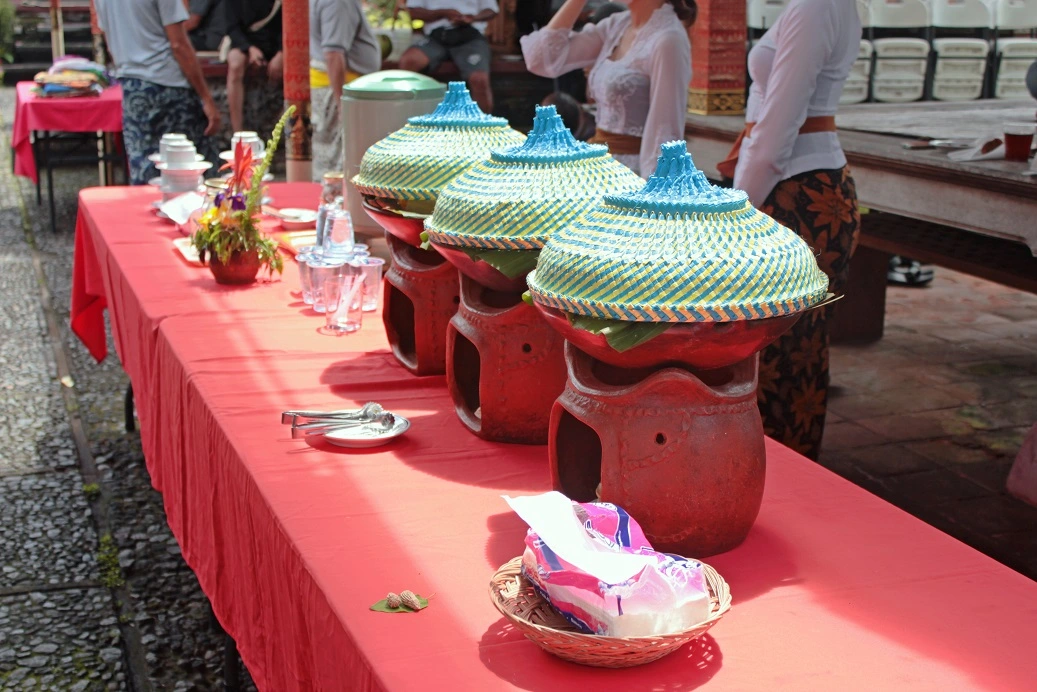 A buffet table set up with traditional Balinese earthenware serving pots covered with colorful woven lids, arranged in a row on a red tablecloth, offering refreshments and lunch to participants after an eco cycling tour