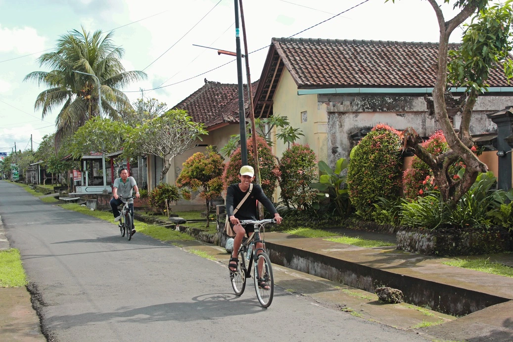 Two individuals partake in an eco cycling tour, riding bicycles down a quiet street lined with traditional Balinese homes and lush tropical plants, under a partly cloudy sky, signifying a serene day of exploration