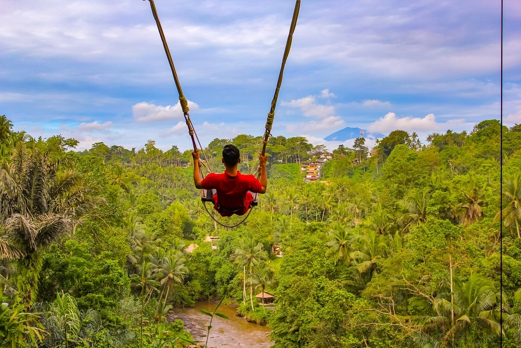 The image captures a breathtaking view of a verdant Balinese landscape. A person, wearing a red shirt, is seen from the back, swinging on a large swing that hangs from towering palms. The swing arcs high above a dense tropical forest, providing a panoramic view of lush greenery, a winding river, traditional huts, and a few scattered buildings. In the distance, under a partly cloudy blue sky, the silhouette of a mountain majestically stands, adding to the serene backdrop. The scene conveys a sense of adventure, freedom, and connection to nature.