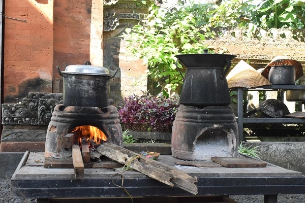 Traditional Balinese clay stoves with pots on top are fired up with wood, cooking outdoors on a stone platform, with intricate Balinese sculptures and lush plants in the background, illustrating a scene of local culinary practices