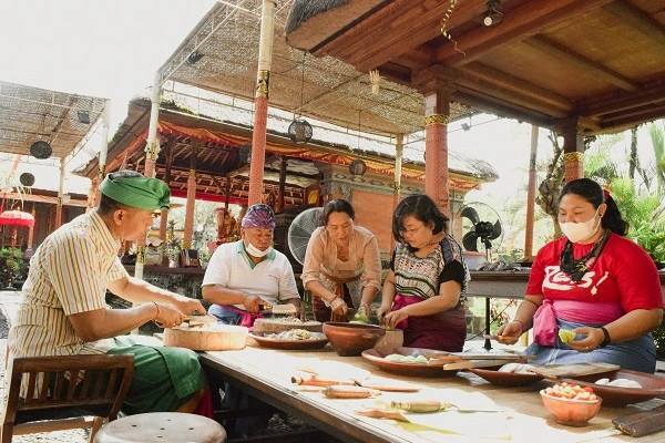 A cooking class in action, with participants gathered around a wooden table, attentively preparing ingredients under the guidance of instructors in a traditional open-air Balinese pavilion